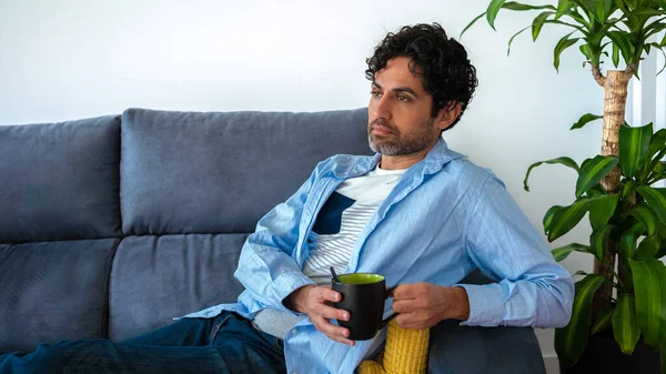 Face of unhappy lonely adult man looking up pensive while drinking morning expresso coffee and sitting alone on couch home. A sad person with a cafe and lost in thoughts at sofa