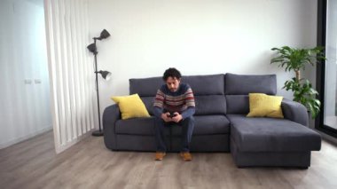 Evdeki kanepede otururken akıllı telefonunu kullanan neşeli yakışıklı bir adam. Kendine güveni tam sakallı adam akıllı telefonuyla evin kanepesinde daktilo çekiyor..