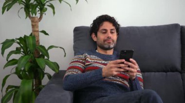 Evdeki kanepede otururken akıllı telefonunu kullanan neşeli yakışıklı bir adam. Kendine güveni tam sakallı adam akıllı telefonuyla evin kanepesinde daktilo çekiyor..