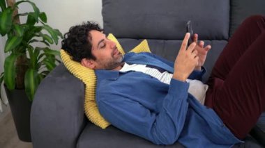 Akıllı telefon kullanırken gülümseyen mutlu beyaz adamın portresi ve güzel bir iç mekanda kanepede uzanmak. Kafkasyalı bir adam telefon cihazıyla ev kanepesinde dinleniyor..