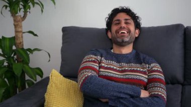 Sakallı, kışlık süveter giyen, gülümseyen, kameraya bakan ve evinin kanepesinde oturan yakışıklı genç bir adam. Pozitif kişi.