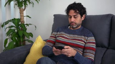 Akıllı telefon kullanırken gülümseyen mutlu beyaz adamın portresi ve güzel bir iç mekanda kanepede oturması. Kafkasyalı bir adam telefon cihazıyla ev kanepesinde dinleniyor..