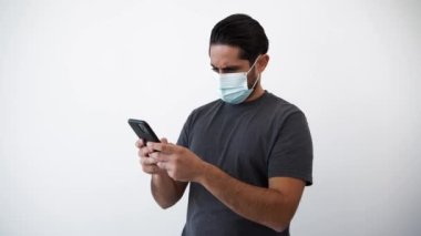 Ciddi ve sinirli bir adam beyaz arka planda izole edilmiş koronavirüsten korunmak için akıllı telefon ve yüz maskesi kullanıyor. Negatif bir insan elinde bir telefon cihazı tutuyor. Covid-19 pandemik karantina kavramı