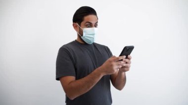 Beyaz adamın portresi, Corona virüsü salgını sırasında akıllı telefon ve tıbbi maskeyle sürpriz yaparak kaşlarını kaldırıyor. Beyaz arka planda telefon cihazı olan şaşırmış tepki veren kişi.
