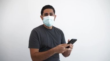 Akıllı telefon ve tıbbi maske kullanan neşeli beyaz bir adamın portresi Corona virüsü salgını sırasında elde yükseltilmiş bir başarıyla kutlanıyor. Beyaz arka planda izole edilmiş kapalı stüdyo görüntüleri