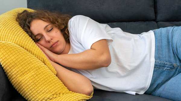 Sleeping woman with curly hair taking nap on the sofa during the day at cozy apartment. Attractive girl sleep lying on the comfortable couch with eyes closed. Dreaming and resting concept