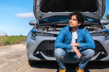 Stresli bir adam yolun kenarında kapüşonlu bir araba arızasından sonra oturuyor. Çekici servisinin yolda kaza yapmasını bekliyor..