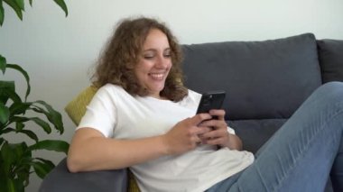 Beyaz kadın akıllı telefon kullanıyor. Haftasonu evdeki kanepede uzanıyor. Mobil uygulama ve cihaz yaşam tarzı. İnternet konseptine göz atan telefon kullanıcıları.
