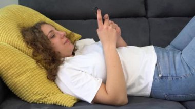 Kıvırcık saçlı bir kadın, rahat bir dairede kanepede otururken akıllı telefon kullanıyor. Kız arkadaş internetten alışveriş yapıyor ve sosyal medya izliyor. İnternet konseptine göz atan telefon kullanıcıları.