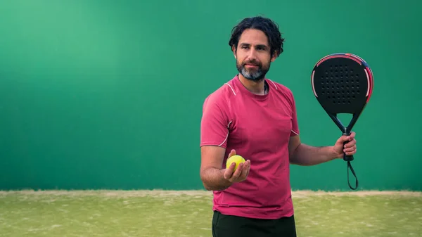 Monitor of padel holding black racket with yellow tennis ball in the hand. Class to student on outdoor tennis court. Man paddel player playing a match in the open