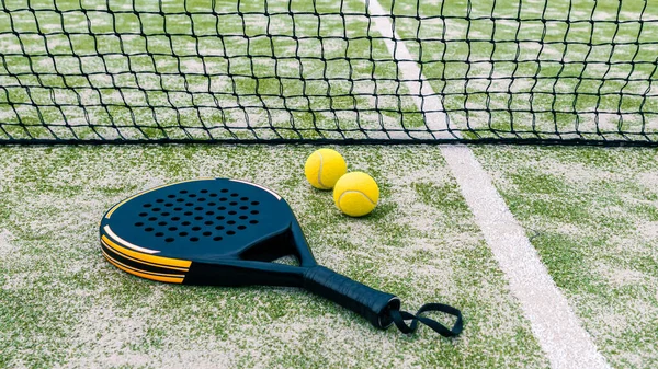 Yellow balls on grass turf near padel tennis racket behind net in green court outdoors with natural lighting. Paddle is a racquet game. Professional sport concept with copy space