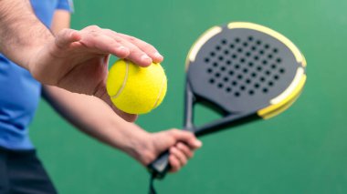 Padel 'in sarı topu ve siyah raketi açık havada tutan profesyonel monitörü. Açık tenis kortunda sınıftan öğrenciye. Adam raketçi açıkta maç yapıyor. Raket sporu konsepti.