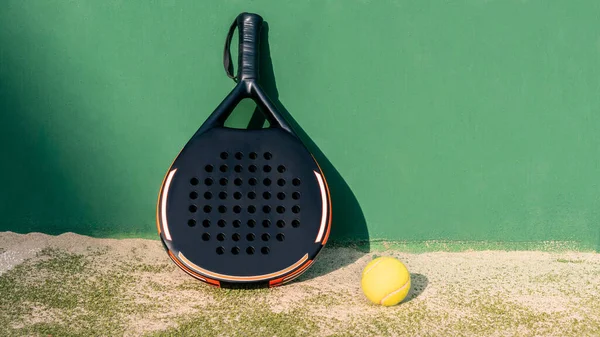 Yellow ball on floor near of padel tennis racket in green court outdoors with natural lighting. Paddle is a racquet game. Professional sport concept with copy space