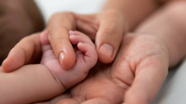 Young mother holding baby hand and showing finger and fingernail on white sheet bed. Closeup of baby sleeping on the mother's room. Family and home concept