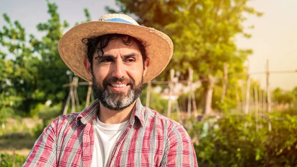 Portrait of a modern bearded farmer man take a break with hoe on hands stands in the agricultural field. Male worker in agricultural farm. Organic vegetables and agriculture farming.