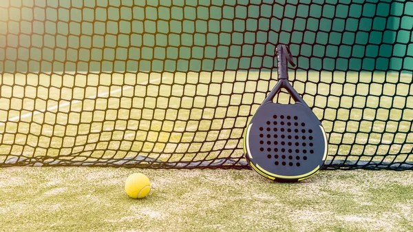 Padel racket and yellow ball behind net on a green court grass turf outdoors at sunset. Paddle is a racquet game. Professional sport concept with space for text