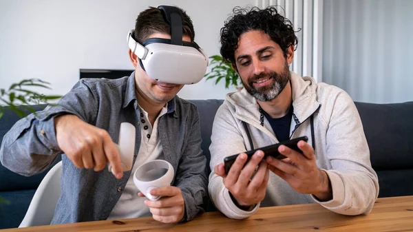 Caucasian men enjoying virtual reality glasses while sitting at the table in home. Two happy guys gesturing and using vr headset or 3d spectacles playing together video game
