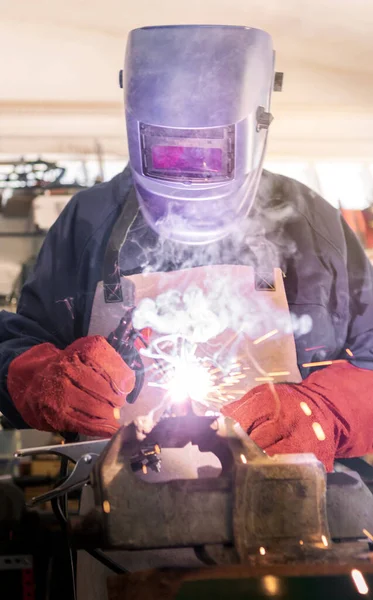 Worker in special clothes and helmet works as welding machine in his workshop, bright sparks from metal heating and fly in different directions. The man works with an electric tool