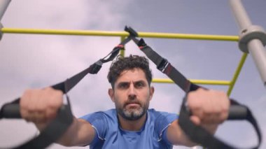 Young man doing fitness exercises on a special hanging device. Fitness male guy doing exercising with suspension trainer slingin a city park. A suspended TRX sport fitness concept.