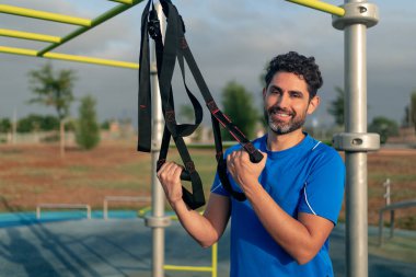 Portrait of confident caucasian man during workout with suspension TRX on the street. Young active guy does suspension training with fitness straps outdoors in city park.