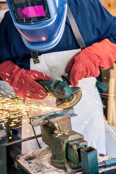 Worker in special clothes and goggles grinding metal with a circular saw in his workshop, bright sparks of metalworking fly in different directions.