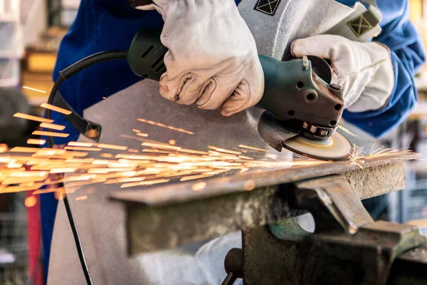 Worker in special clothes and goggles grinding metal with a circular saw in his workshop, bright sparks of metalworking fly in different directions.