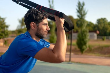 Young man doing fitness exercises on a special hanging device. Fitness male guy doing exercising with suspension trainer slingin a city park. A suspended TRX sport fitness concept.