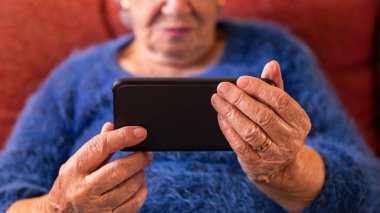 Evde cep telefonu kullanan yaşlı bir kadın. Büyükannenin buruşuk elleri akıllı telefon tutuyor. Teknoloji kavramı