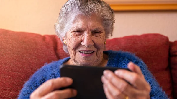 Elderly woman using a mobile phone while sitting on sofa home. Happy smiling grandmother holds a smartphone. Old grandma operating a telephone. Concept of technology device