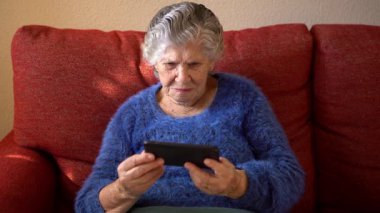 Kanepede otururken cep telefonu kullanan yaşlı bir kadın. Büyükannemin akıllı telefonu var. Yaşlı büyükanne telefon kullanıyor. Teknoloji aygıtı kavramı