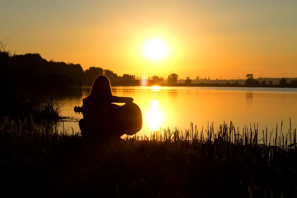 Silhouette di una ragazza all'alba che suona la chitarra vicino al fiume — Foto Stock