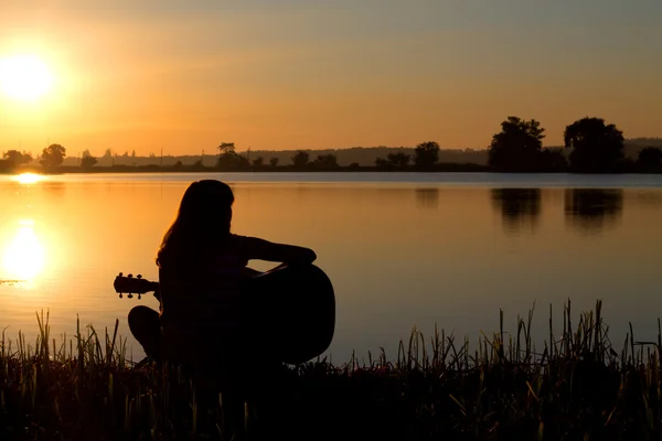 Sziluettje egy lány gitározni a folyó naplementekor — Stock Fotó