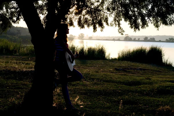 Silueta de una chica tocando una guitarra cerca de un árbol — Foto de Stock