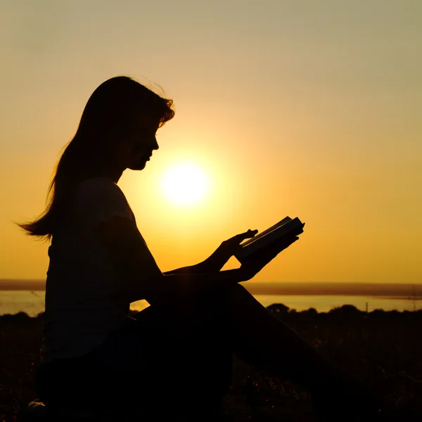 Silhueta de uma menina com um livro nas mãos ao pôr do sol — Fotografia de Stock