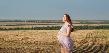 Güzel, kızıl saçlı, hamile bir kadın bir buket kır çiçeğiyle tarlada dikiliyor ve bulutlu gökyüzüne bakıyor.