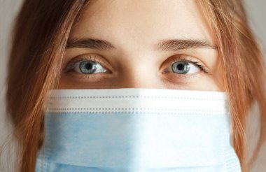 Genç, kızıl saçlı, mavi gözlü, koruyucu bir maske takan, gri stüdyo geçmişi olan, hamile bir kadının mutlu yüzü, sağlık ve ilaç koruma kavramı.