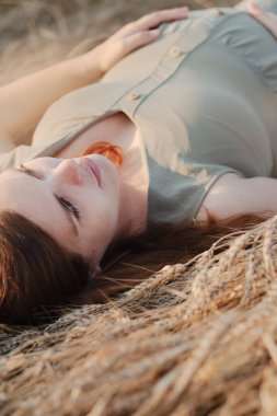 Beyaz hamile, genç, kızıl saçlı bir kadın yazın gün batımında buğday tarlasında uzanıyor, müstakbel anne doğada dinleniyor, sağlıklı ve mutlu hamilelik kavramı, yeni bir hayat.