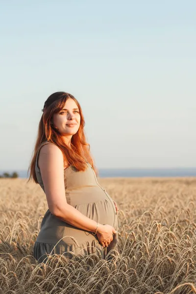 美丽的年轻孕妇在日落时走在麦田上 红头发的孕妇在大自然中轻松自在地用手抚摸着她的肚子 快乐的怀孕观念 — 图库照片