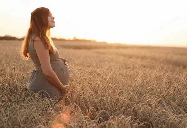 美丽的年轻孕妇在麦田的夕阳西下抬起头来 红头发的准妈妈在大自然中轻抚着她的肚子 快乐的怀孕观念 — 图库照片
