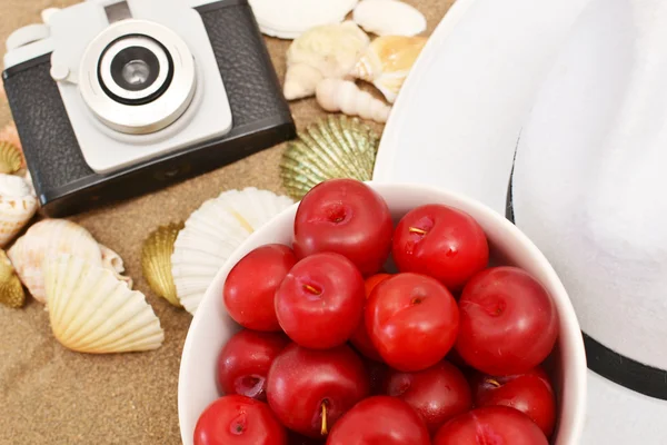 Red plums, old camera, shells and white hat on sand — Stock Photo, Image