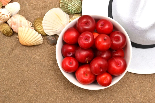 Prunes rouges, coquillages et chapeau blanc sur sable — Photo