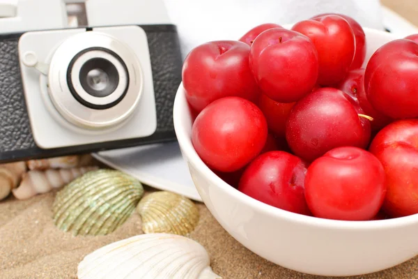 Red plums, old camera, shells and white hat on sand — Stock Photo, Image