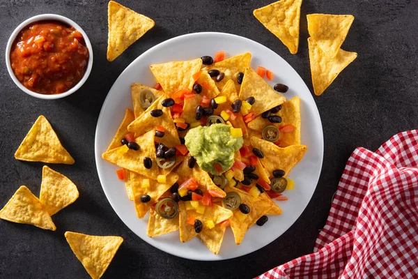 Mexican nachos tortilla chips with black beans, guacamole, tomato and jalapeno on black background