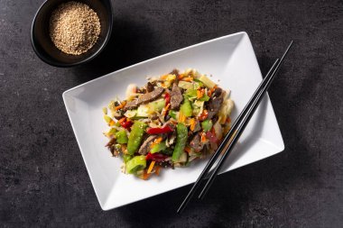 Siyah arka planda beyaz tabakta sığır eti, sebze ve susam tohumları.