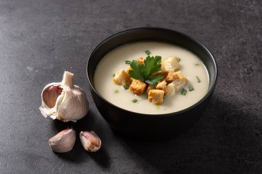 Sarımsak çorbasının üstü kızarmış ekmeklerle kaplanmış siyah kayrak zemin üzerinde.