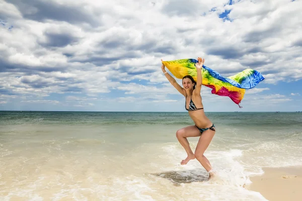 Tanned girl in bikini jumping on the beach with a colored scarf — Stock Photo, Image