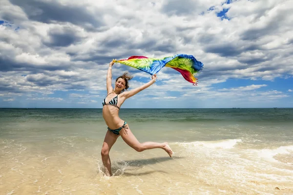 Tanned girl in bikini jumping on the beach with a colored scarf — Stock Photo, Image