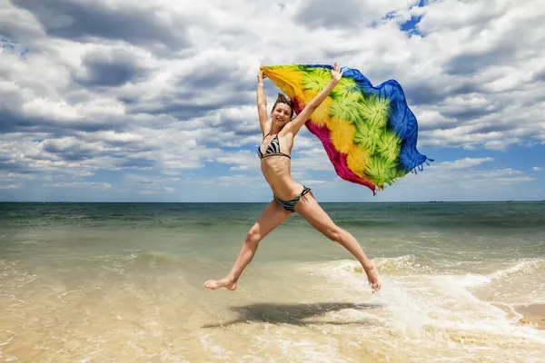 Tanned girl in bikini jumping on the beach with a colored scarf — Stock Photo, Image