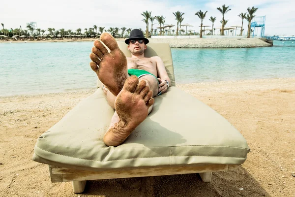 O homem no chapéu banhos de sol em uma cadeira de estar na praia em um — Fotografia de Stock