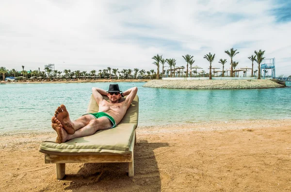 The man in the hat sunbathes on a lounge chair on the beach at a — Stock Photo, Image